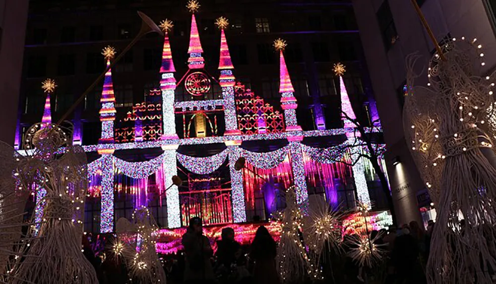 The image shows a spectacular display of holiday lights covering what appears to be a large building set against a night sky with decorative light structures resembling stars or snowflakes in the foreground