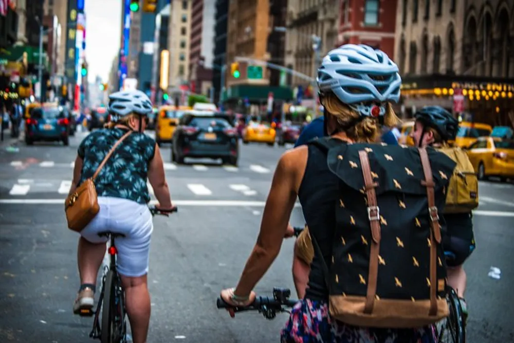 Two cyclists wearing helmets are riding down a bustling city street with taxis and colorful signs in the background