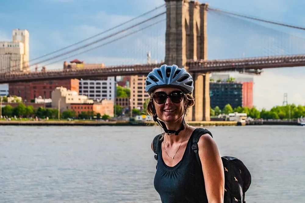 A smiling person with a bicycle helmet stands in front of a scenic view featuring the Brooklyn Bridge and waterfront buildings