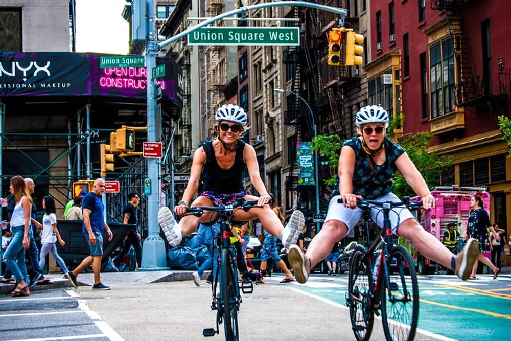 Two cyclists wearing helmets are smiling and riding bikes through an urban street setting with the sign Union Square West overhead