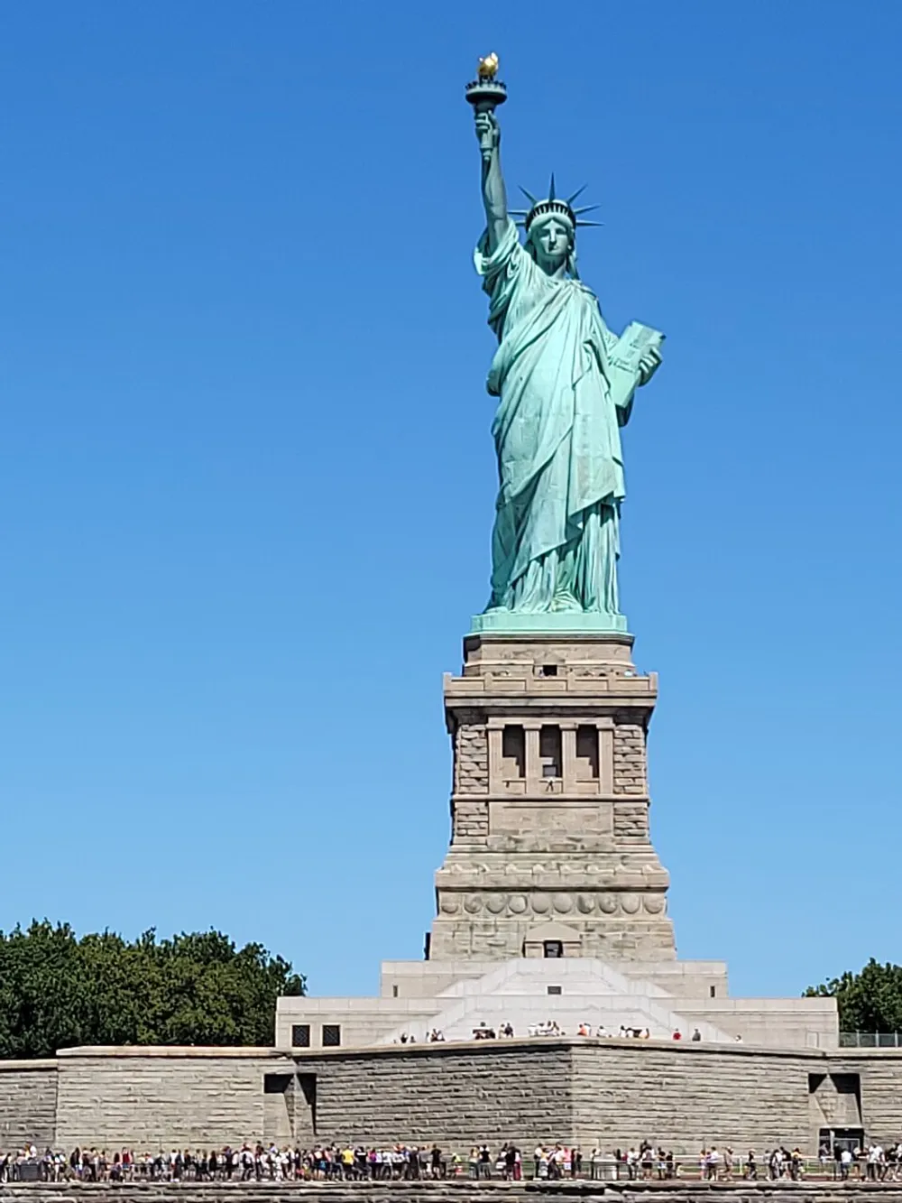 The image shows the Statue of Liberty against a clear blue sky with numerous visitors at its base