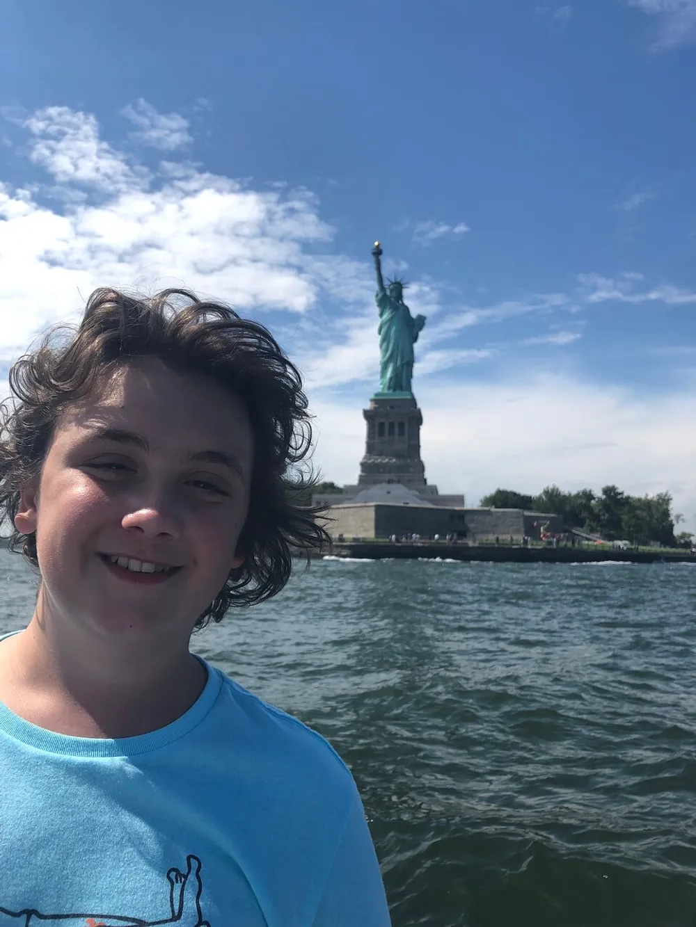 A smiling person is posing in the foreground with the Statue of Liberty visible in the background