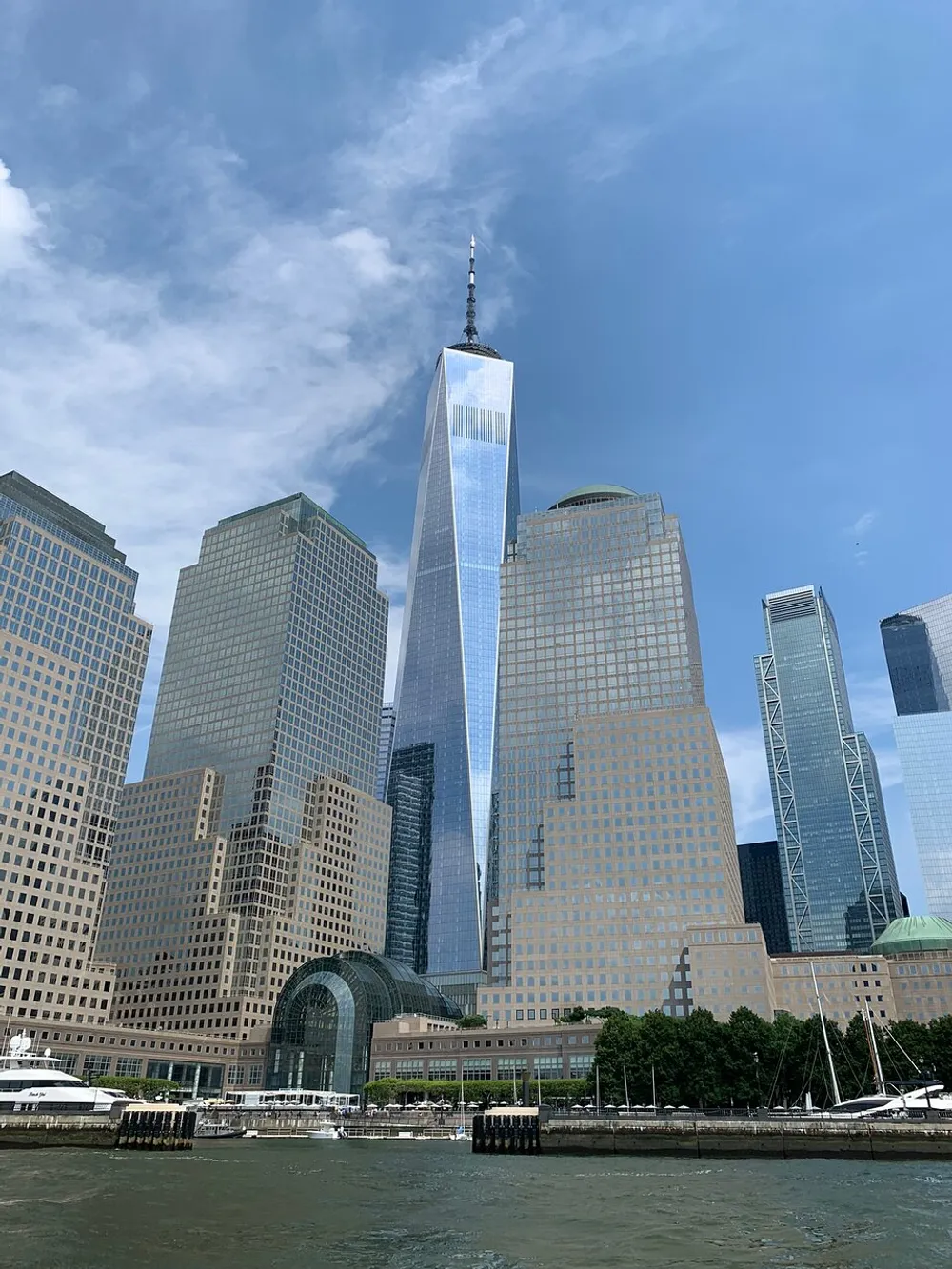 The image shows the skyline with the One World Trade Center dominating the backdrop viewed from the water under a partly cloudy sky