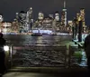 A nighttime view of a brightly lit city skyline seen from a pier with dark waters in the foreground