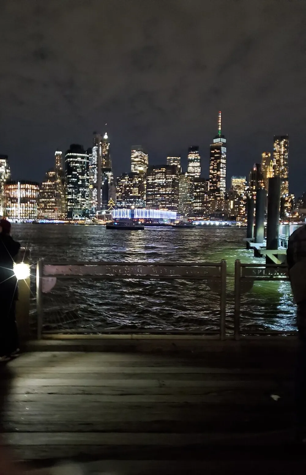 A nighttime view of a brightly lit city skyline seen from a pier with dark waters in the foreground