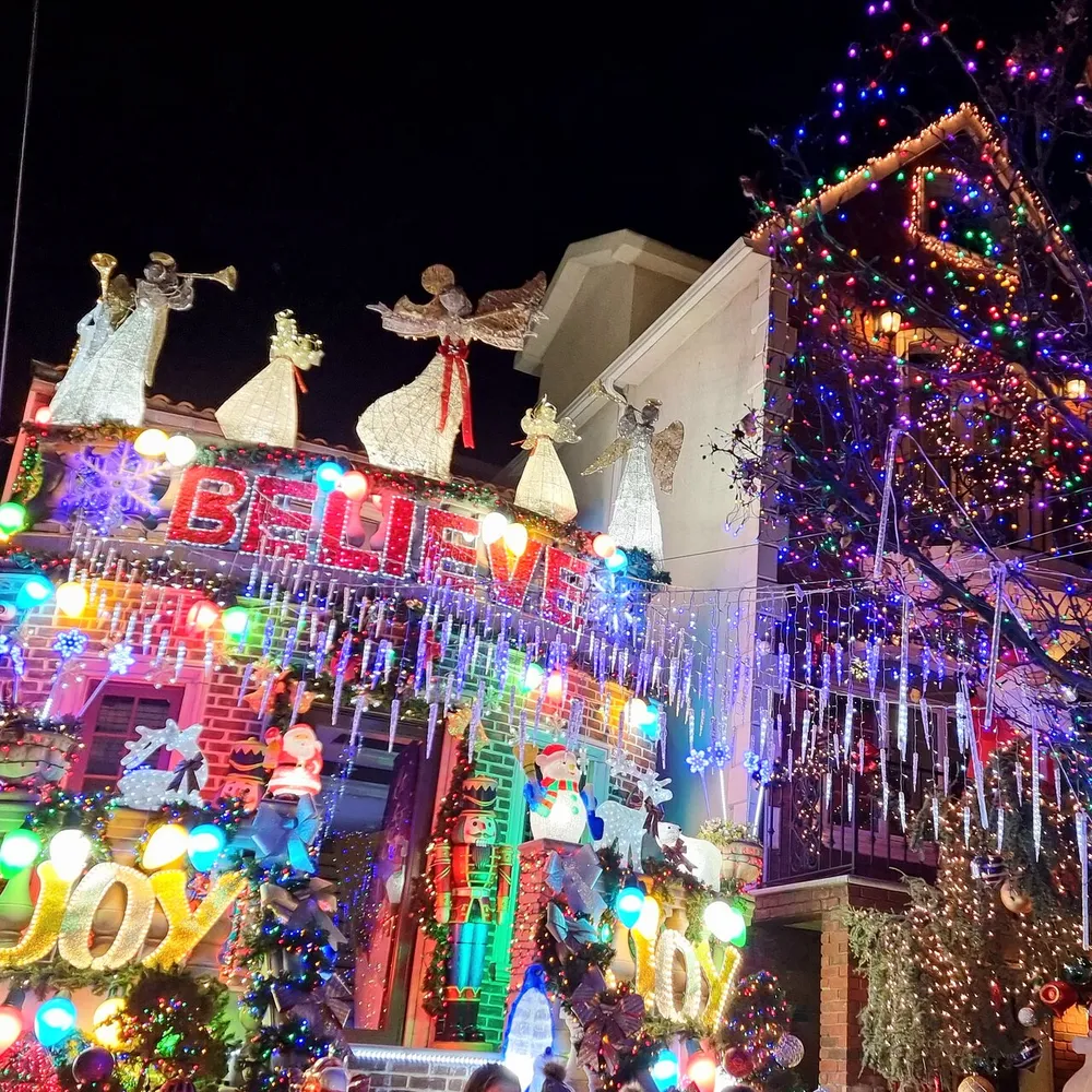 The image shows a vividly decorated house with bright Christmas lights festive figures and words like BELIEVE and JOY conveying a highly spirited holiday atmosphere