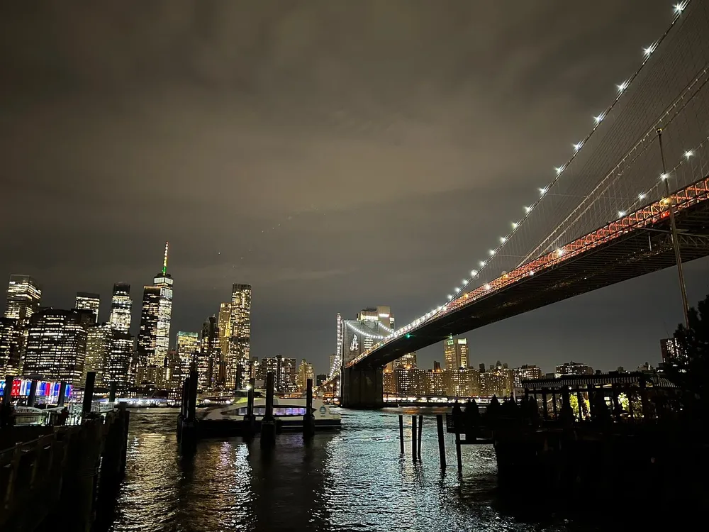 The photo captures a night scene featuring a brightly lit skyline with tall buildings the illuminated Brooklyn Bridge extending over calm waters and a slightly cloudy sky above