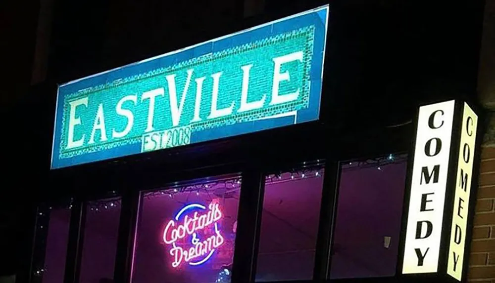 The image features the illuminated signage of EastVille Comedy Club at night advertising stand-up comedy with a prominent blue EASTVILLE sign and a vertical COMEDY marquee