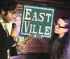 A man holding a microphone speaks to a woman on a stage with a sign that reads EASTVILLE Est 2008 in the background