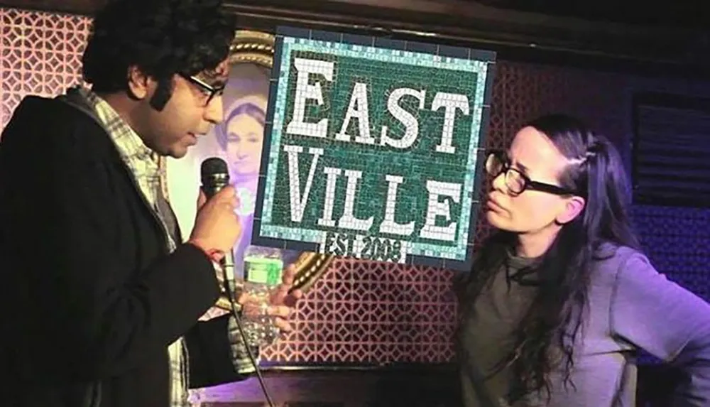 A man holding a microphone speaks to a woman on a stage with a sign that reads EASTVILLE Est 2008 in the background