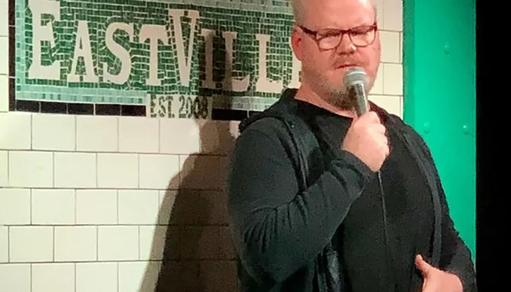 A person is performing stand-up comedy at a club called EastVille as indicated by the mosaic sign in the background