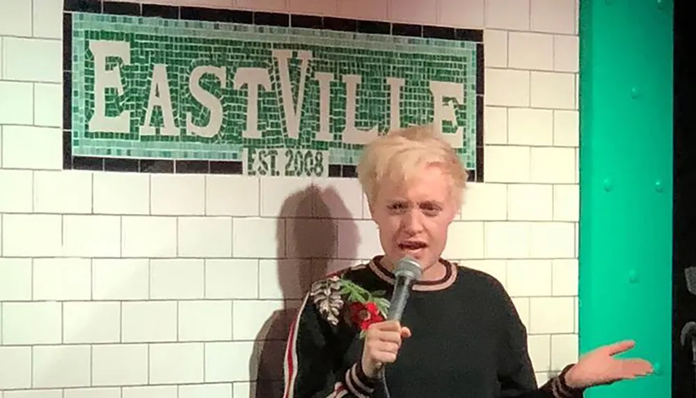 A person is performing stand-up comedy at a venue with EASTVILLE est 2008 on the wall behind them