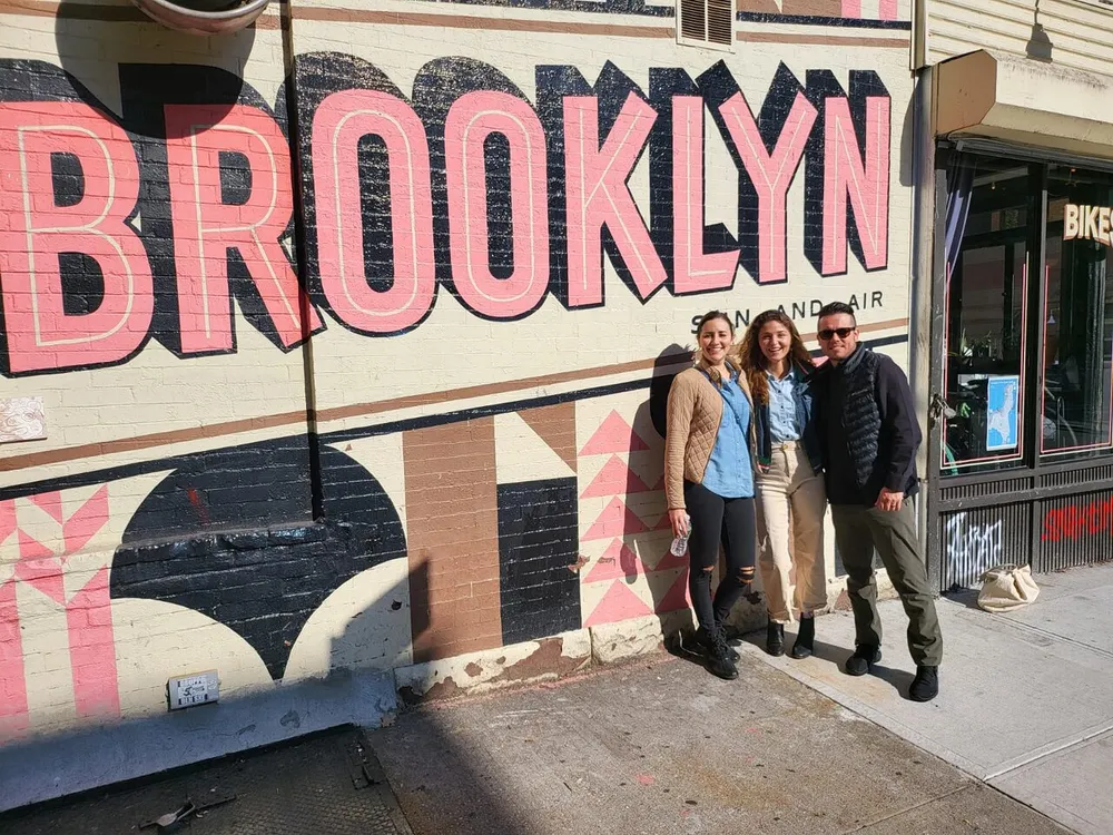 Three people are smiling and posing in front of a large mural that spells out BROOKLYN on a sunny day