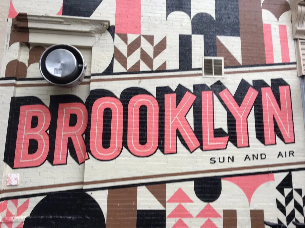 The image shows a large bold mural with the word BROOKLYN painted on a buildings wall accompanied by the phrase SUN AND AIR and geometric decorative elements