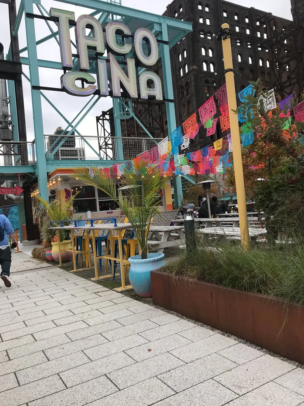 The image shows an outdoor seating area of a colorful Mexican restaurant named Tacocina decorated with bright papel picado banners against the backdrop of an industrial structure