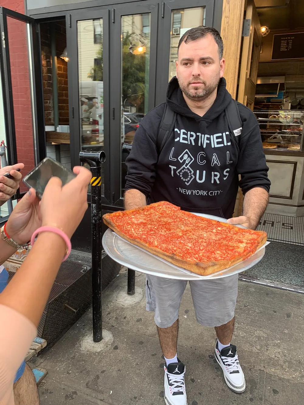 A man is holding a very large slice of pizza on a plate while someones hand is visible with a phone likely taking a photo of the impressive slice