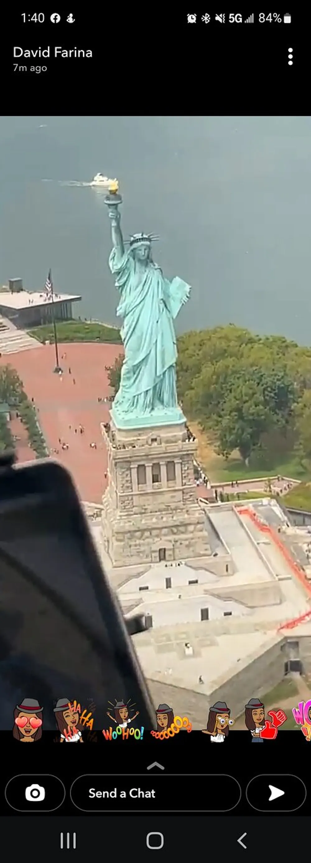 A screenshot from a mobile device displaying an aerial view of the Statue of Liberty with a decorative Snapchat caption and stickers at the bottom