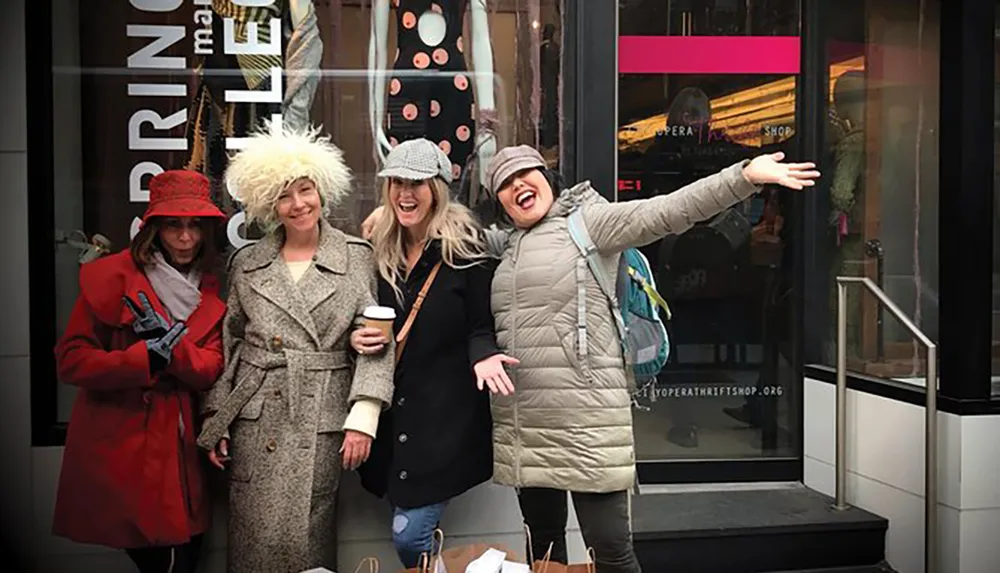Four women are posing for a photo on a city street each expressing different levels of enthusiasm with shopping bags in hand standing in front of a store that has a Bring In Spring Sale sign