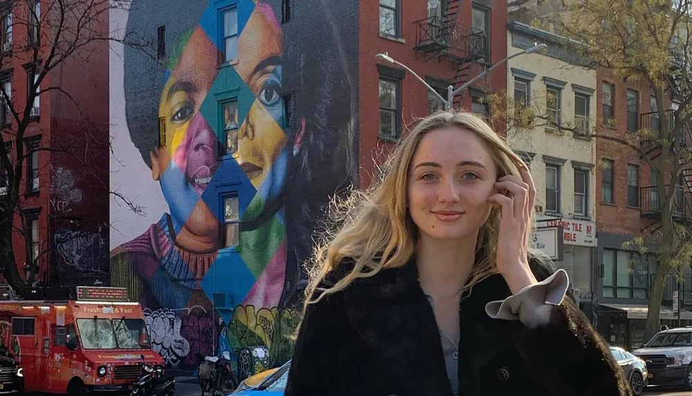 A woman with blond hair is smiling in the foreground with a vibrant mural of a persons face depicted in various colors in the background on a city street