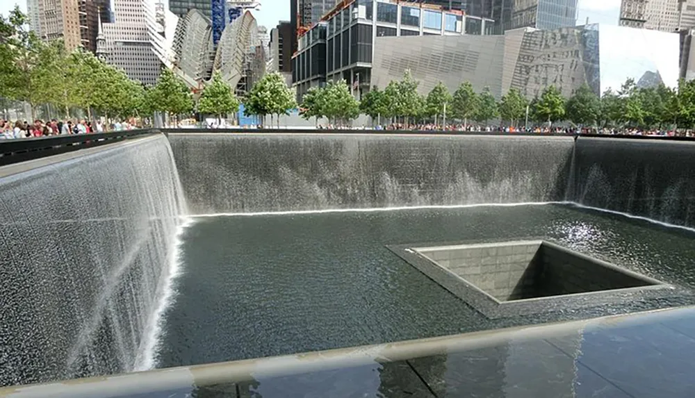 The image shows a memorial pool with cascading waterfalls surrounded by a plaza with visitors set against a backdrop of modern buildings