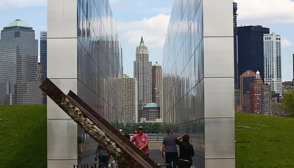 The image shows visitors at a reflective memorial with a weathered metal beam in the foreground and a cityscape in the background