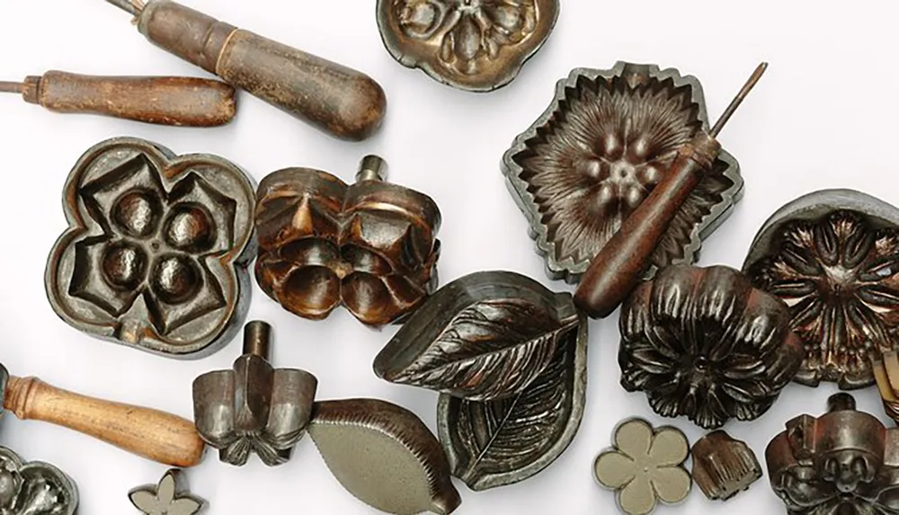 The image shows a collection of antique metal cookie molds of various floral and leaf designs accompanied by wooden-handled tools possibly used for making detailed confectionery