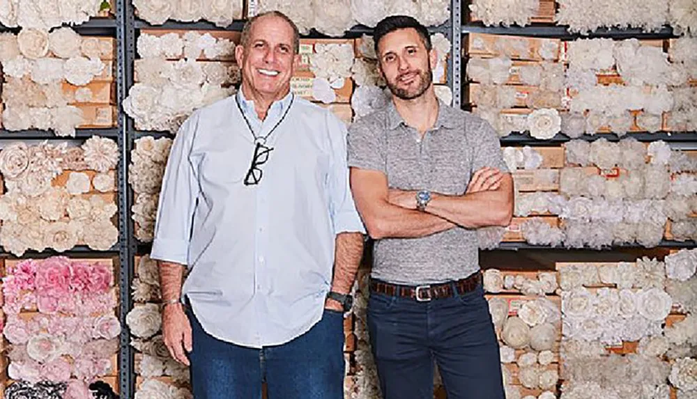 Two men are standing in front of shelves filled with assorted decorative flowers smiling for the camera