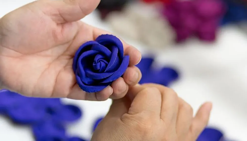 A person is holding a blue fabric rose in their hand against a blurred background of colorful materials