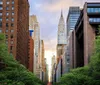 The image depicts a lush tree-lined street in a bustling city with the iconic Chrysler Building standing tall amidst surrounding skyscrapers against a backdrop of a dramatic evening sky