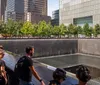 The image captures visitors observing a memorial with cascading waterfalls on the footprint of a former building surrounded by trees and modern skyscrapers in an urban setting