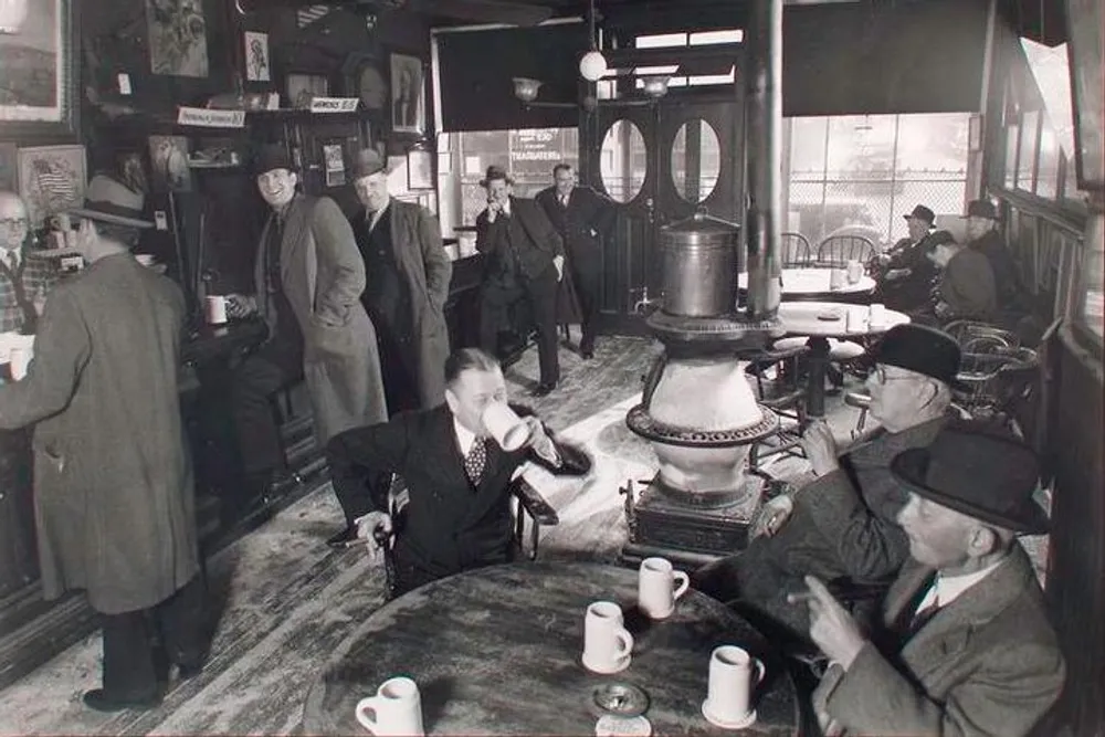 The black-and-white image captures a moment inside a bustling vintage tavern or cafe where patrons clad in mid-20th-century attire engage in conversation drink and warm themselves by a large stove