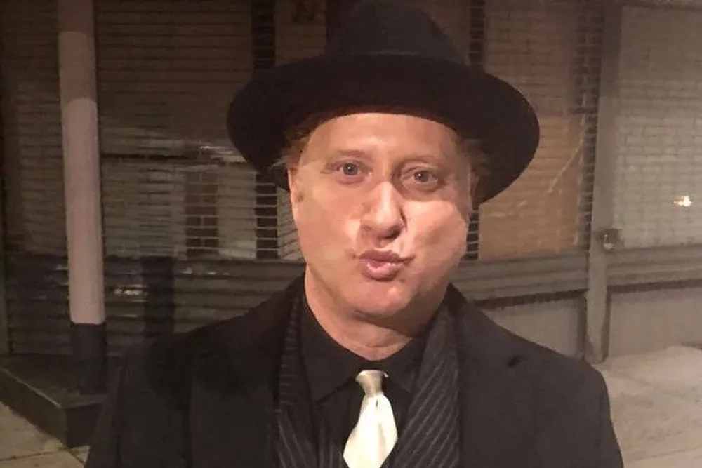 A person is making a puckered lip expression while wearing a black hat suit and tie standing against an urban backdrop at night