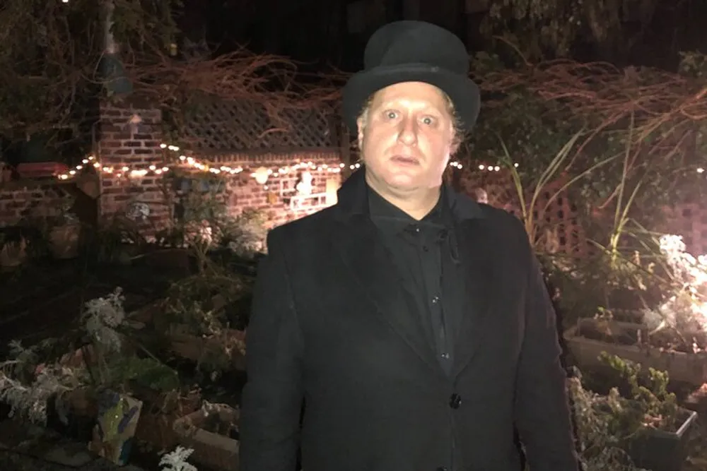 A person wearing a black coat and top hat is standing in front of a garden with twinkling lights at night