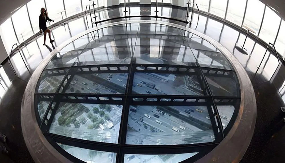 A person stands on a transparent glass floor inside a high-rise building providing a vertiginous view of the street and vehicles far below