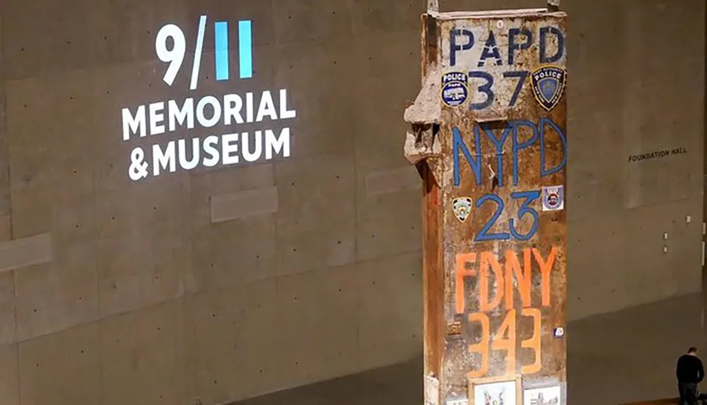 The image shows a section of a destroyed column with inscriptions and emblems standing as a remembrance inside the 911 Memorial  Museum with a person observing it