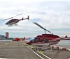 A helicopter is preparing for takeoff on a helipad near a body of water while another helicopter flies overhead and a third one is parked in the background