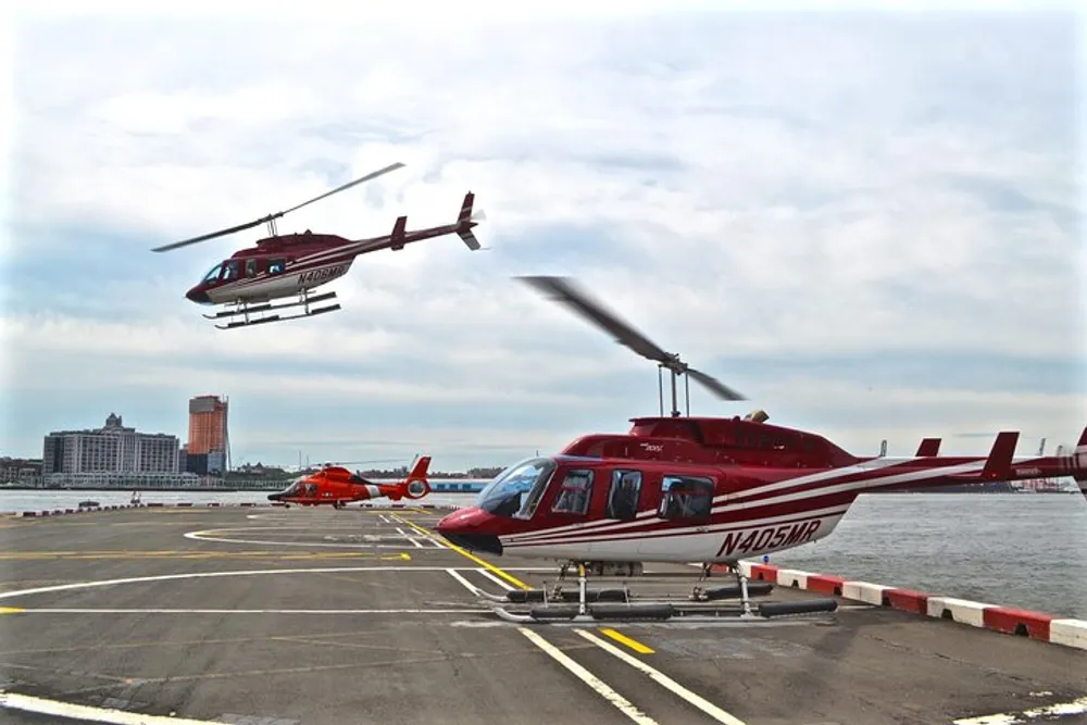 A helicopter is preparing for takeoff on a helipad near a body of water while another helicopter flies overhead and a third one is parked in the background