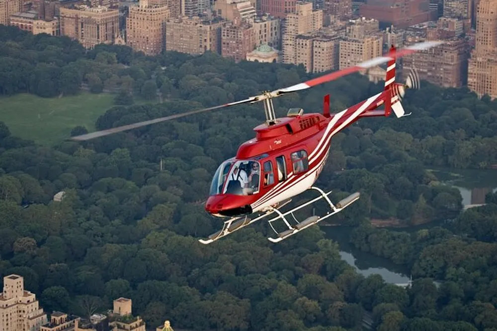 A red helicopter is flying over a large green park surrounded by city buildings