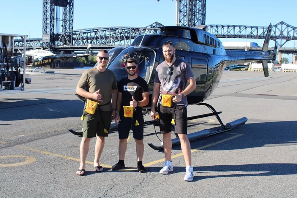 Three individuals are standing and smiling in front of a helicopter each wearing similar yellow safety equipment