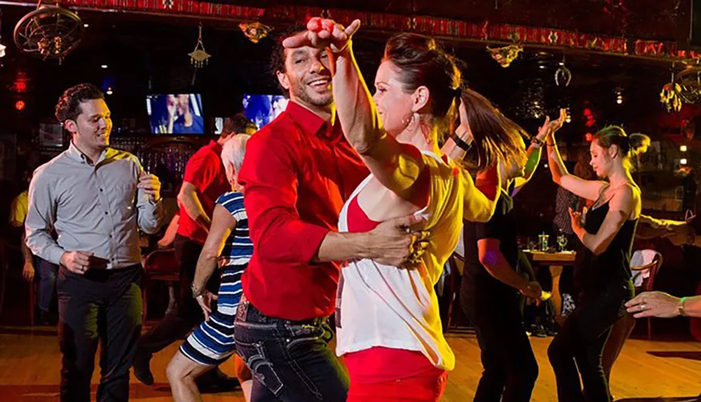 A group of people are joyfully engaging in social dancing at a vibrant venue with a couple in the foreground performing a dance move with eye contact and smiles