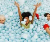 Three people are joyfully submerged in a sea of blue plastic balls reminiscent of a ball pit raising their arms and smiling