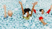 Three people are joyfully submerged in a sea of blue plastic balls, reminiscent of a ball pit, raising their arms and smiling.