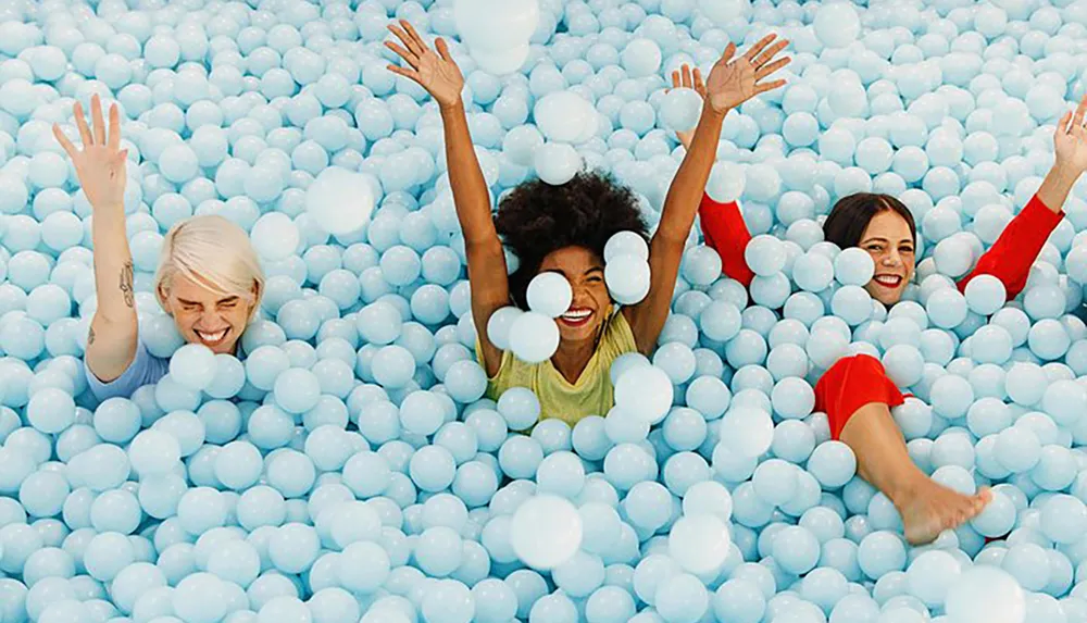 Three people are joyfully submerged in a sea of blue plastic balls reminiscent of a ball pit raising their arms and smiling