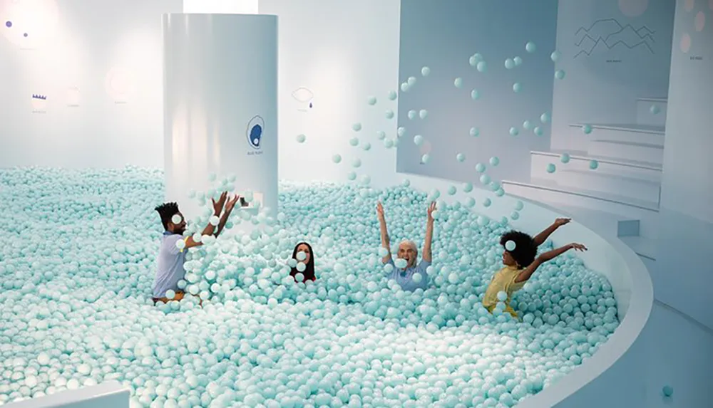 The image shows a playful indoor scene where multiple people are having fun in a large pit filled with small light blue balls reminiscent of a ball pit typically found in childrens play areas