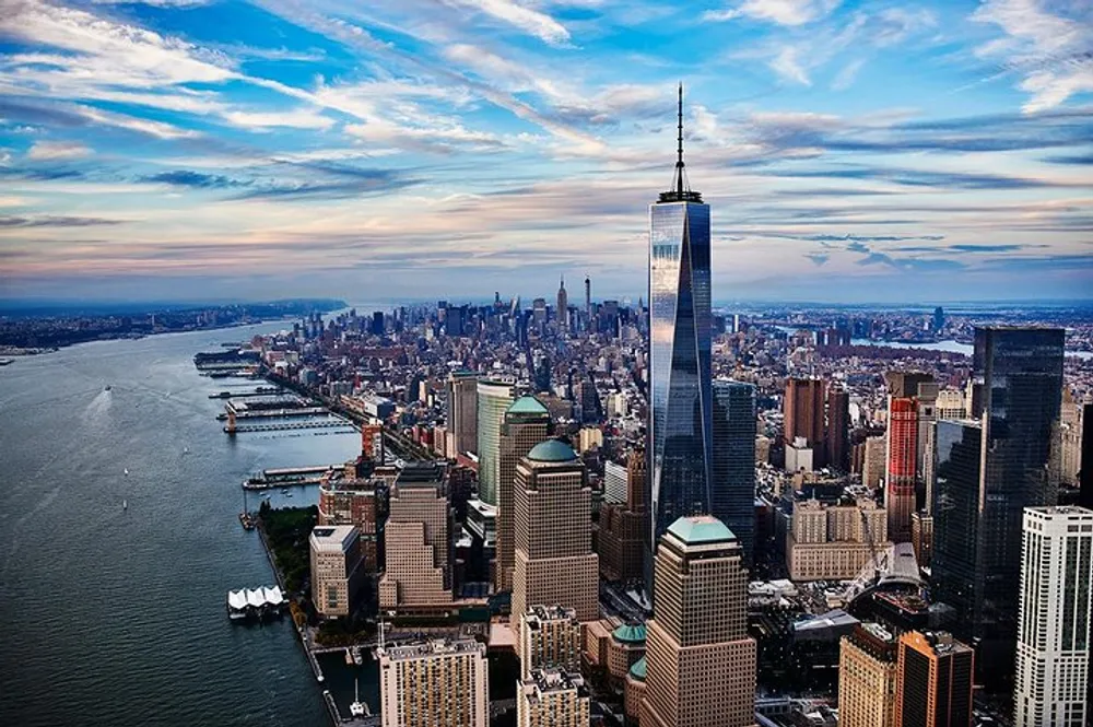 An aerial view of Lower Manhattans skyline dominated by a tall skyscraper near a broad river under a partly cloudy sky