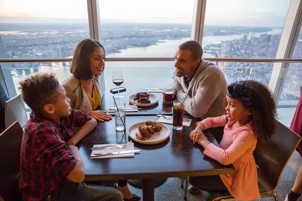 A family of four enjoys a meal together at a high-rise restaurant with a panoramic view of a cityscape in the background