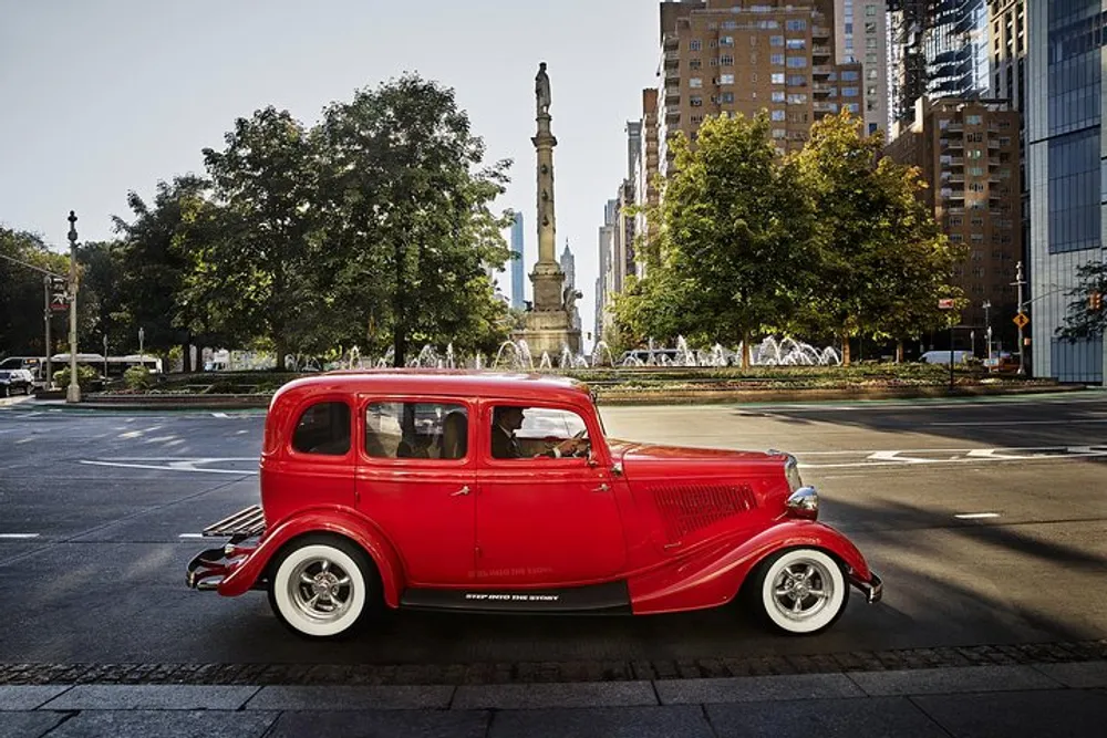 A vibrant red vintage car is parked on a city street with a fountain and a monument in the background under a clear sky