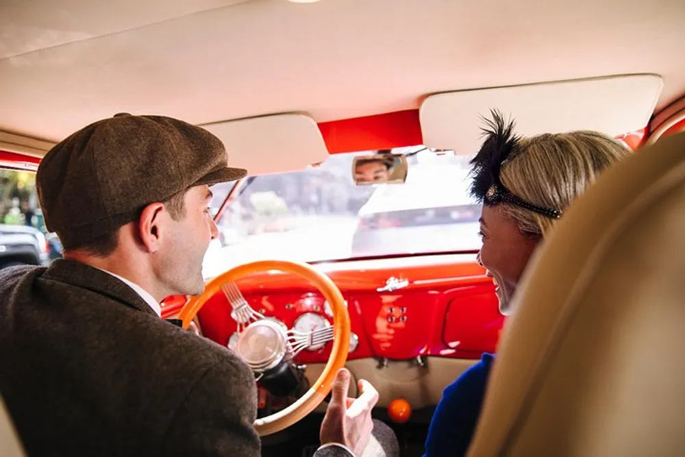Two people dressed in vintage clothing are having a cheerful conversation inside a classic car with a bright red interior