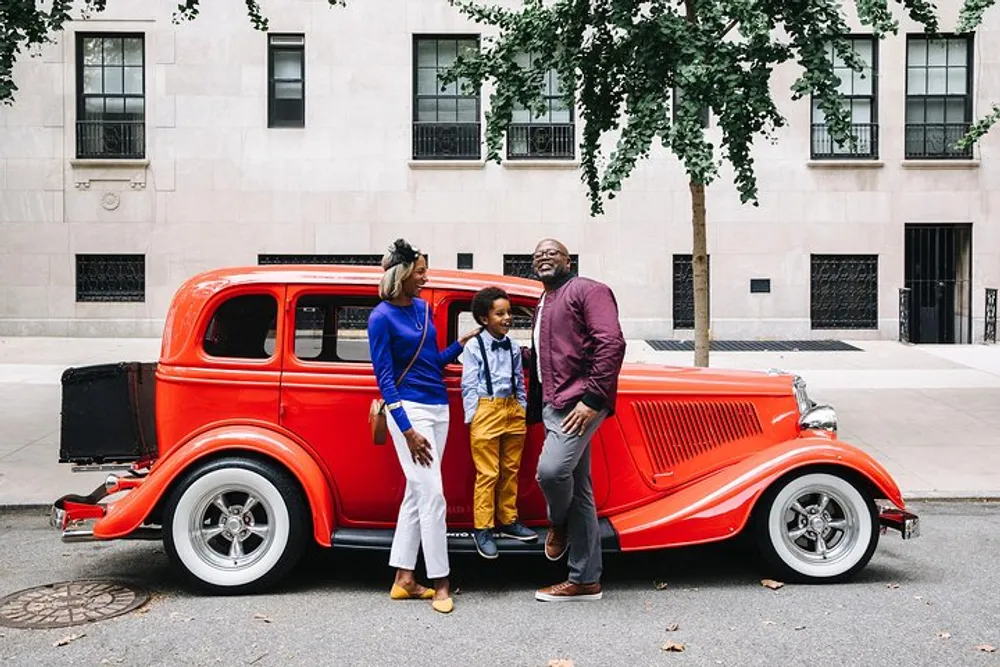 A family of three is smiling and posing with a vintage red car on an urban street