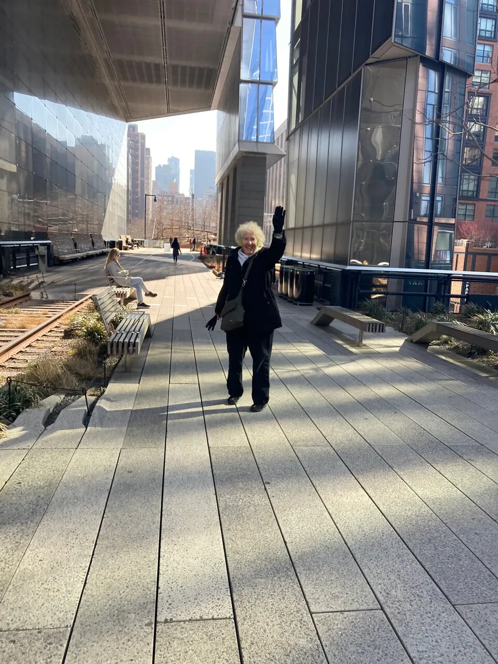 A person with light hair waves cheerfully while walking along an urban highline park with high-rise buildings and seated individuals in the background